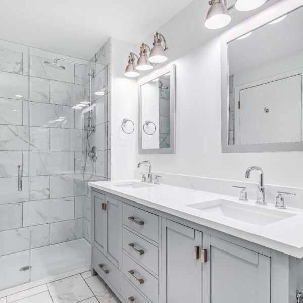 An elegant, remodeled bathroom with a grey vanity and bronze hardware. The shower has a large shower head and marble tiles and glass wall line the sides.